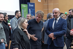 Kilka grup osób zgromadzonych podczas swobodnych rozmów w sali wykładowej. Na pierwszym planie grupa trzech osób, od lewej strony kobieta i mężczyzna ubrani na czarno i mężczyzna w ciemnym garniturze.