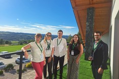 Pięć osób stoi obok siebie na balkonie i pozuje do zdjęcia na tle naturalnego letniego krajobrazu. Wszyscy są elegancko ubrani i uśmiechają się. W tle zielone wzgórza porośnięte lasami.