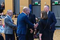 Na sali wykładowej grupa pięciu osób, czterech mężczyzn i kobieta podczas przywitania. Wszyscy oficjalnie ubrani. W tle uczestnicy wykładu.