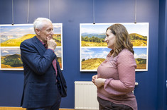 Na pierwszym planie w pomieszczeniu stoją dwie osoby, kobieta w różowej bluzce i mężczyzn w ciemnej marynarce. Są zwróceni do siebie i rozmawiają ze sobą. W tle na granatowej ścianie zawieszone plansze z dużymi kolorowymi fotografiami.