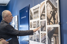 Mężczyzna w ciemnym garniturze wskazuje dłonią na fragment wywieszonego zdjęcia.