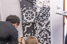 Mężczyzna ubrany na czarno przypina do białej tablicy biało-czarną makietę.