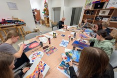 Przy rozległym stole zgromadziło się kilkanaście osób podczas wykonywania swoich prac malarskich. W tle wyposażenia sali plastycznej.