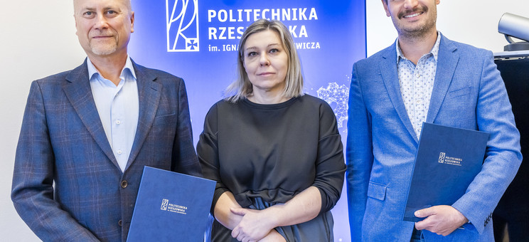 Na tle białej ściany z niebieskim pasem z napisem politechnika rzeszowska stoją obok siebie trzy osoby dwóch mężczyzn i kobieta po środku. Wszyscy elegancko ubrani w różnych odcieniach niebieskiego. Uśmiechają się. Mężczyźni trzymają w rękach okładki z dokumentami.