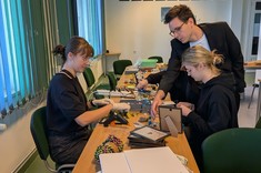 W pomieszczeniu przy długim stole siedzą dwie kobiety i wykonują prace plastyczne. Na stole zgromadzono różne materiały. Z boku mężczyzna nachyla się i wskazuje wybrany materiał. 